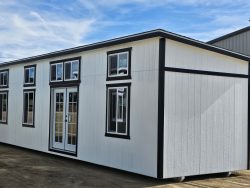 White modular building with black trim and glass doors on an open dirt lot under a blue sky.