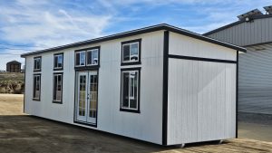 White modular building with black trim and glass doors on an open dirt lot under a blue sky.