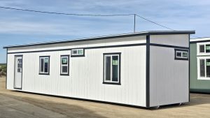 White modular building with black trim and multiple windows on a dirt lot under a blue sky.