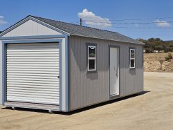 Gray prefab shed on a dirt lot with a large white roll-up door and two small windows under a blue sky.