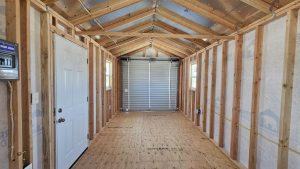 Interior of a framed room under construction with exposed wooden studs, plywood floor, and a metal roll-up garage door at the far end