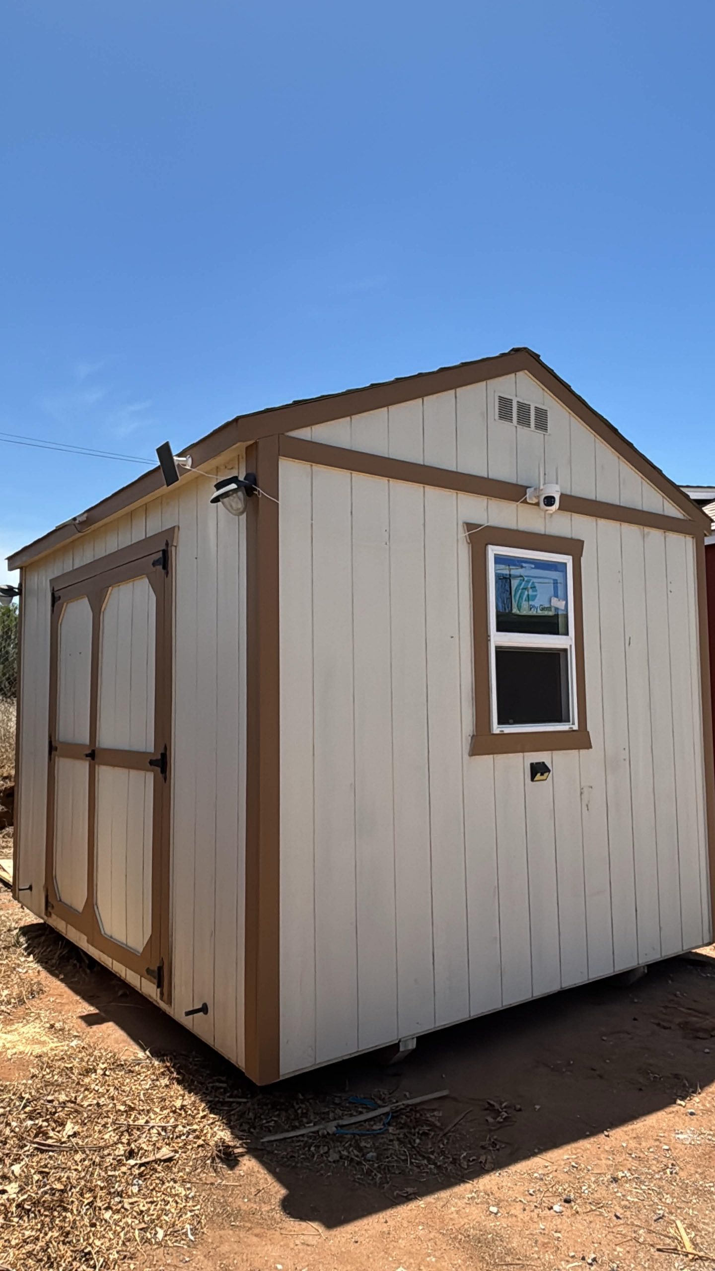Small beige storage shed with brown trim, a window and double doors, under a clear blue sky.
