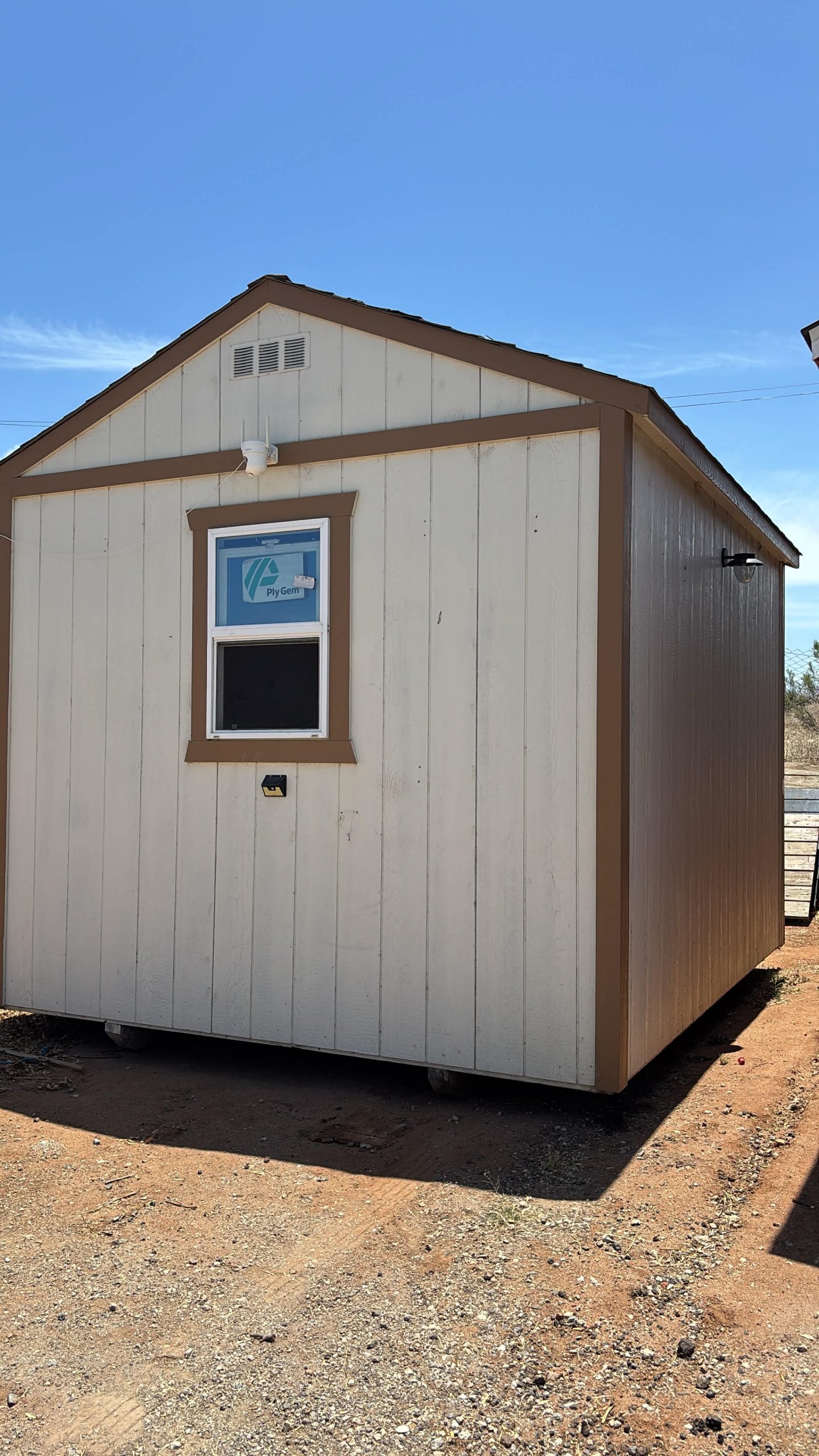 Small beige shed with brown trim under a clear blue sky, featuring a single window and exterior light above it.