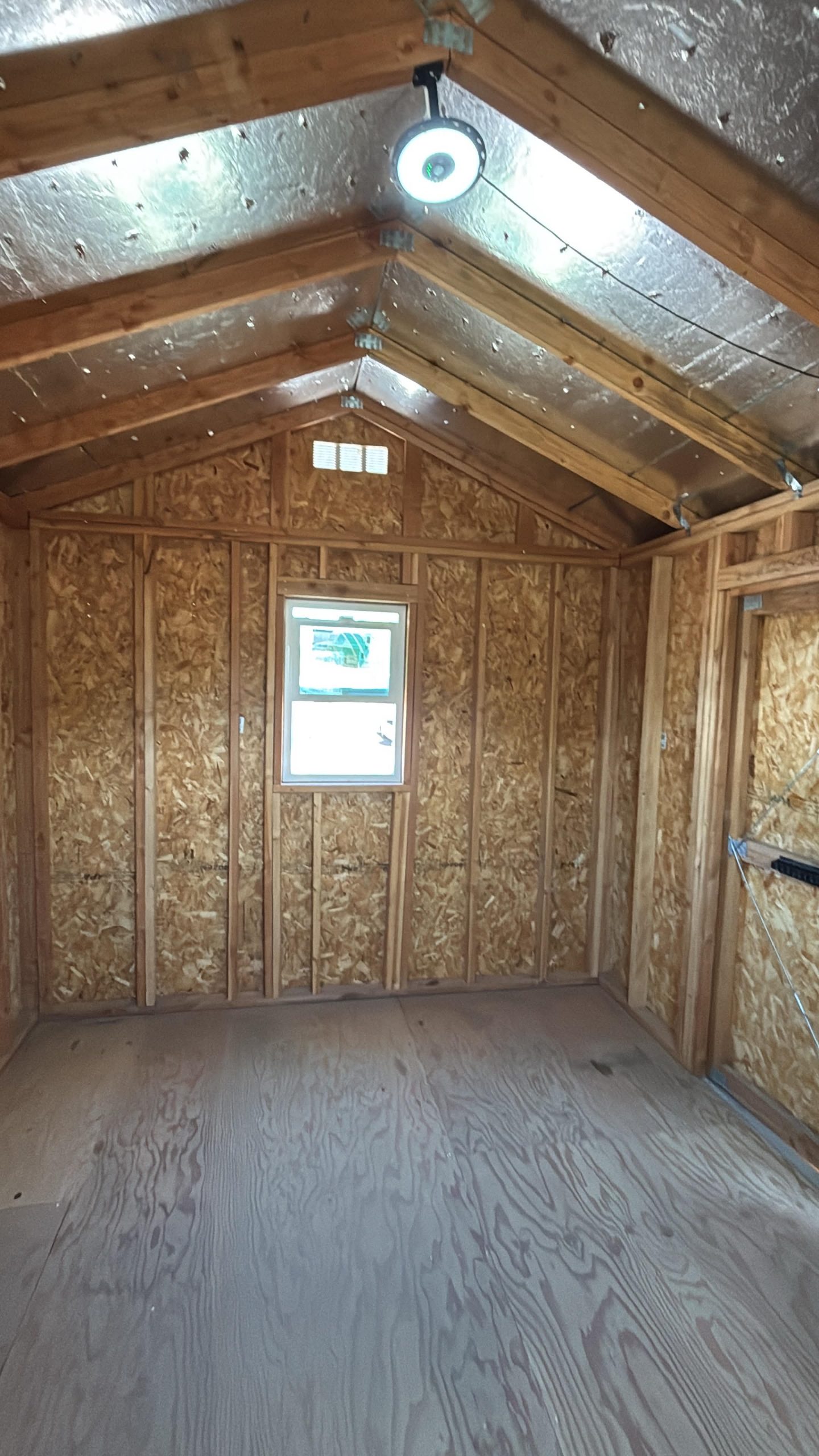 Unfinished attic with wooden framing, OSB walls, a small window, and a round ceiling light with wiring visible.
