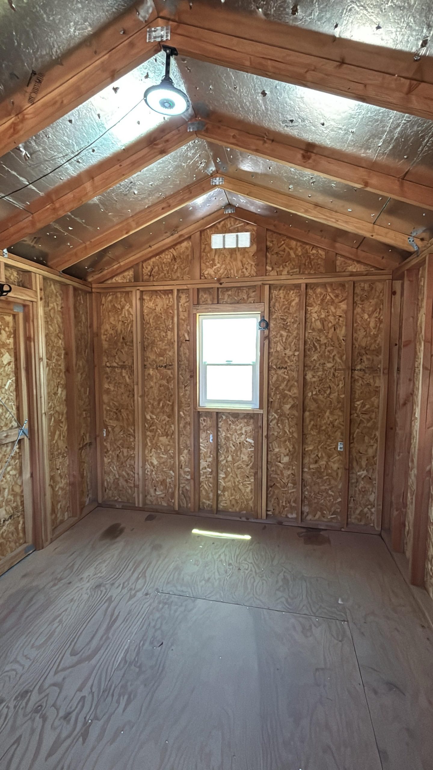 Attic room under construction with exposed wooden framing, foil insulation on the ceiling, a circular hanging light, and a small back wall window.