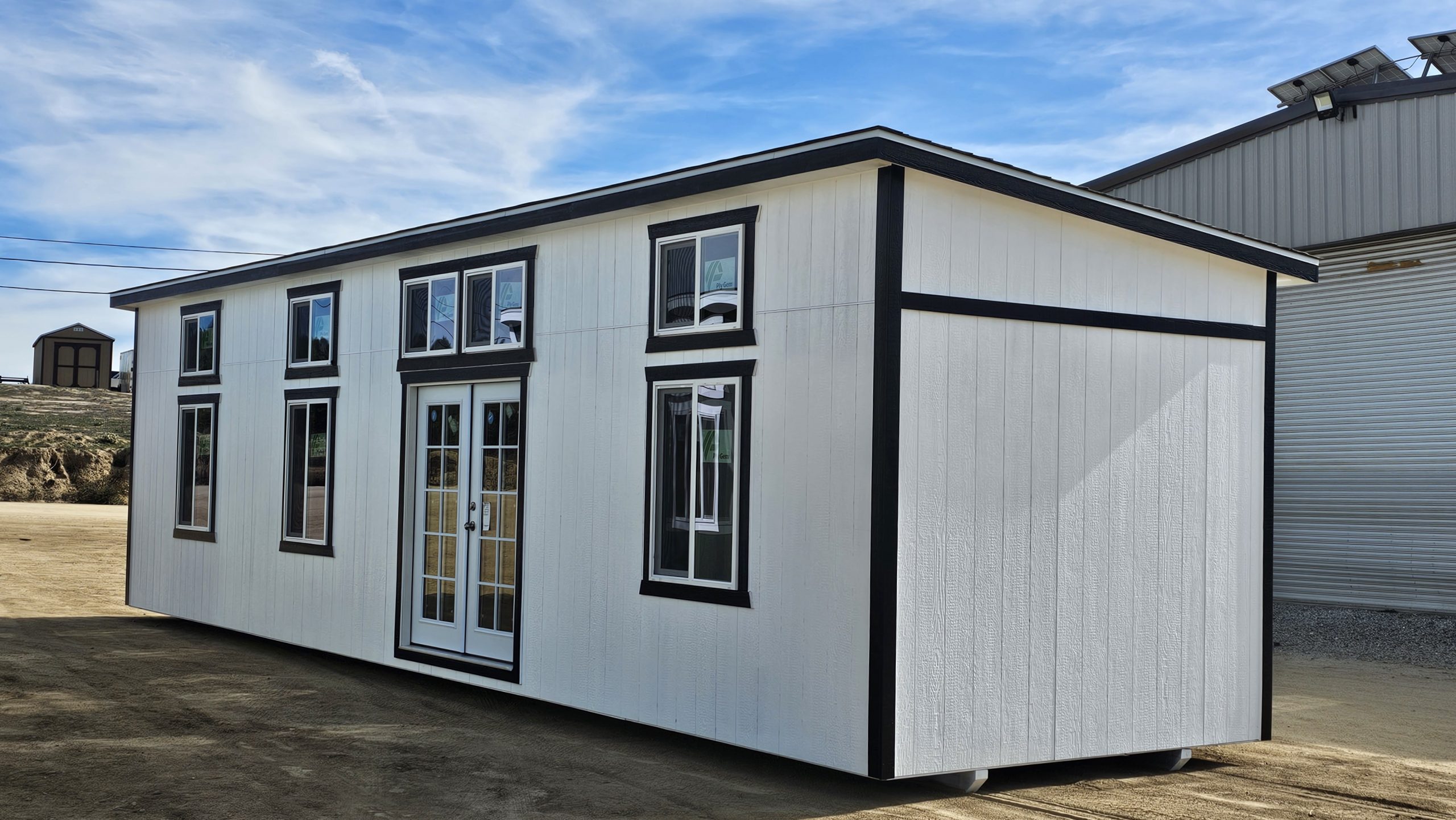 White modular building with black trim and glass doors on an open dirt lot under a blue sky.