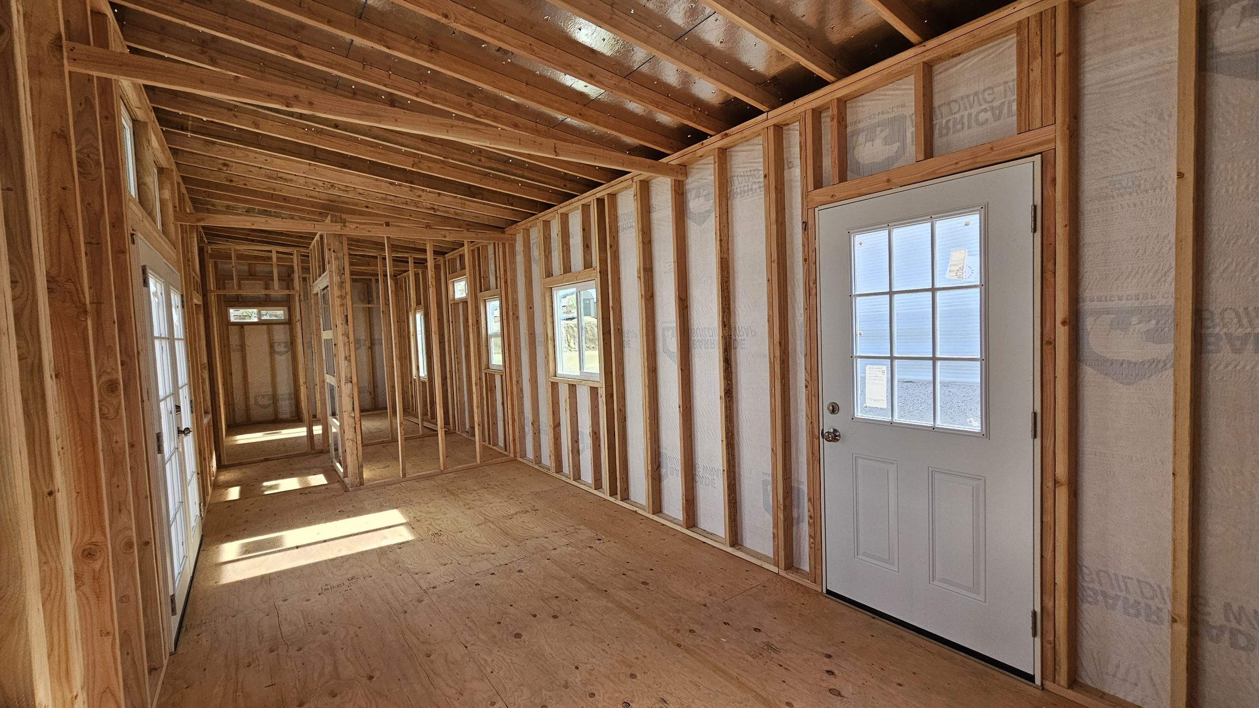 Unfinished interior showing wooden stud framing, exposed ceiling joists, and a white door with glass panes on the right, natural light entering from windows