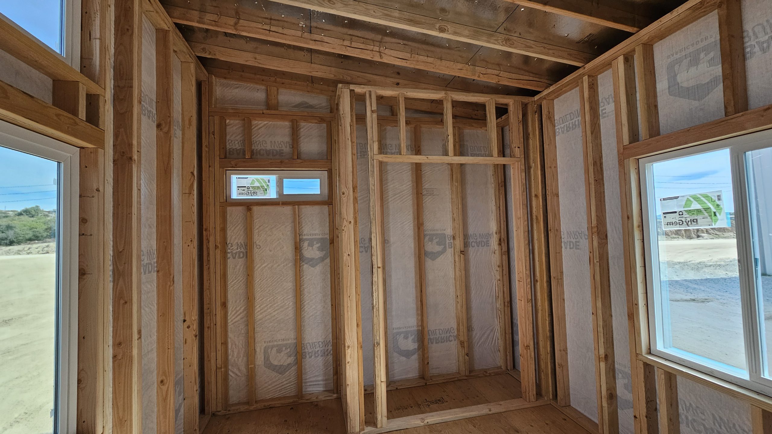 Interior of a new home under construction; exposed wooden studs, ceiling joists, and framed walls with windows letting in daylight.