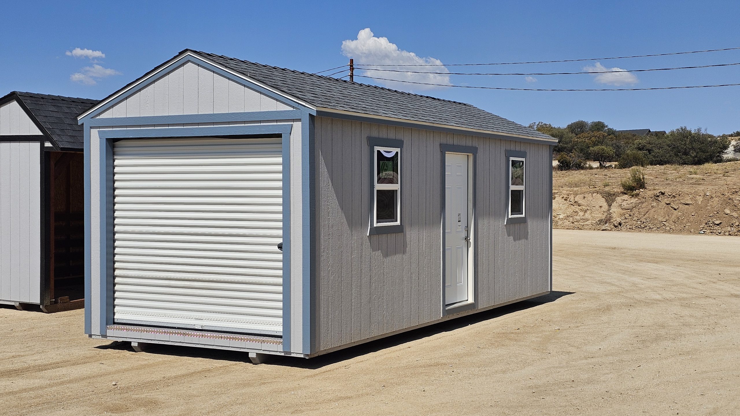 Gray prefab shed on a dirt lot with a large white roll-up door and two small windows under a blue sky.