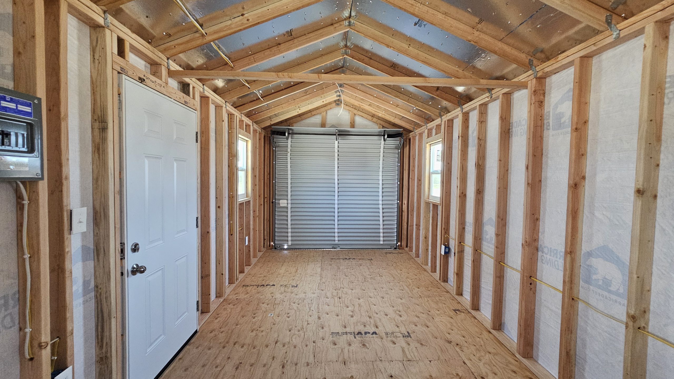 Interior of a framed room under construction with exposed wooden studs, plywood floor, and a metal roll-up garage door at the far end