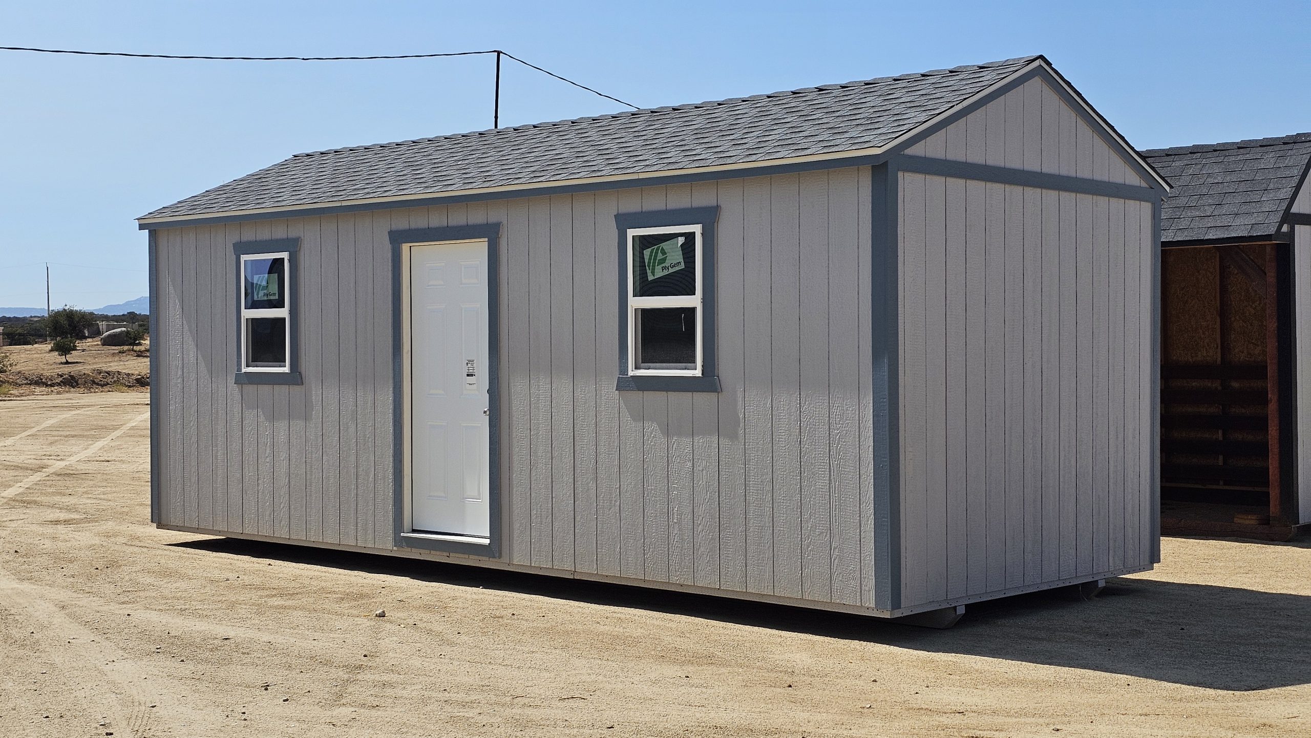 Gray prefab shed with a white door and two small windows on a dirt lot under a bright blue sky, desert landscape in the background.