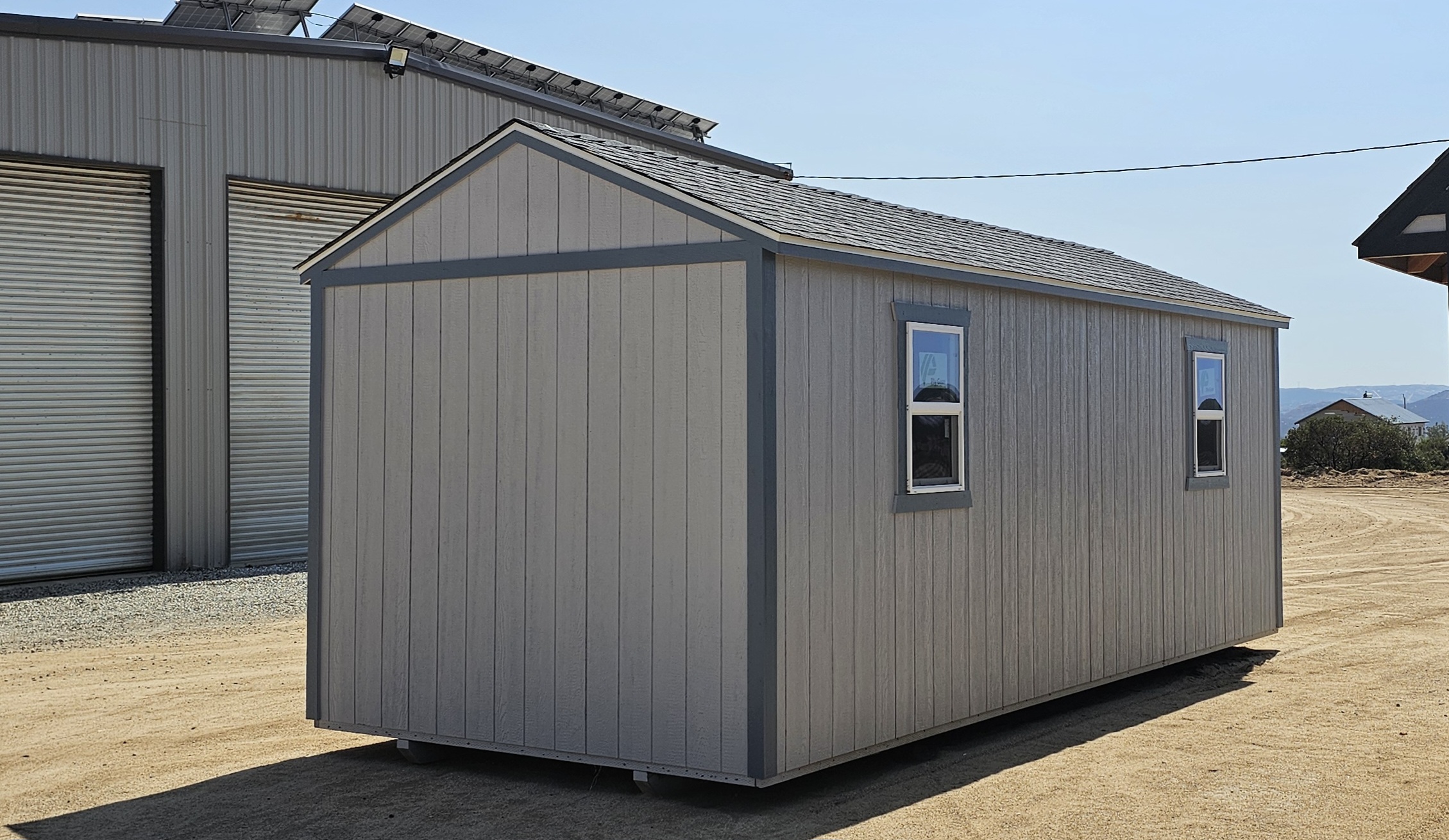 Light-gray portable shed with two windows on its side, set on a dirt lot with a larger metal building in the background.